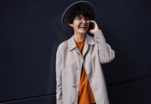 Single Breasted Coat: A Timeless Essential for Modern Men A young person with short curly hair wearing a wide-brim hat, glasses, an orange sweater, and a light beige coat. They are smiling and talking on a smartphone while standing against a dark wall.