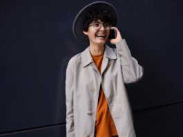 A young person with short curly hair wearing a wide-brim hat, glasses, an orange sweater, and a light beige coat. They are smiling and talking on a smartphone while standing against a dark wall.