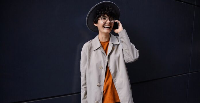A young person with short curly hair wearing a wide-brim hat, glasses, an orange sweater, and a light beige coat. They are smiling and talking on a smartphone while standing against a dark wall.