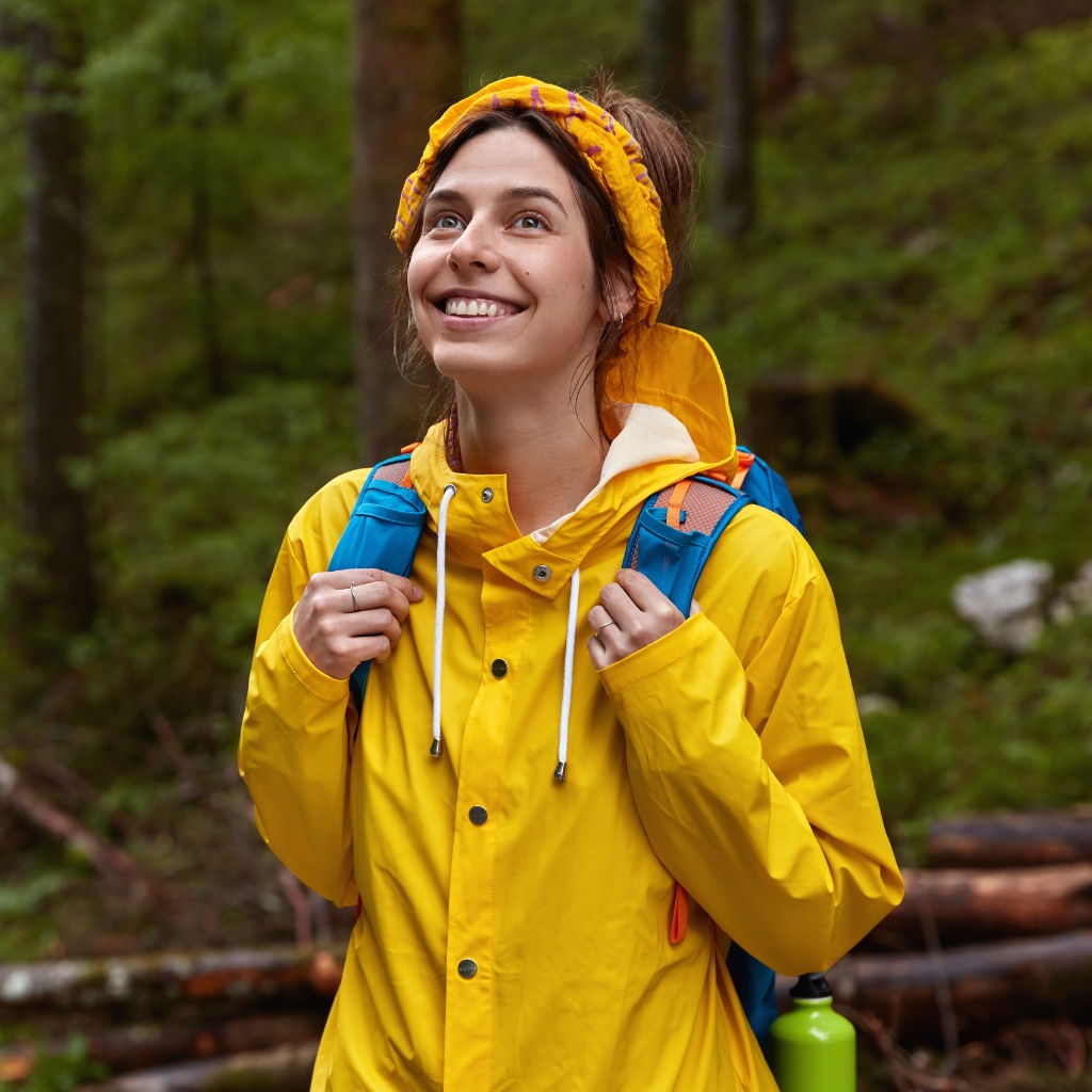  A smiling woman with brown hair wearing a bright yellow rain jacket and a blue backpack stands outdoors in a forest. She has a yellow headband and looks up toward the sky; a green water bottle is visible at the bottom right.
