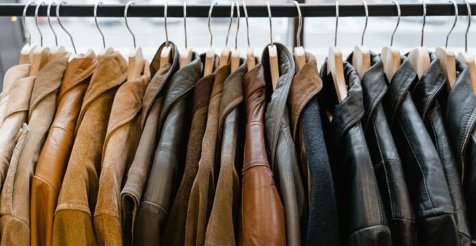 A dense row of leather and suede jackets on a clothing rack, hung on wooden hangers from a black rod. The jackets come in a range of brown and black tones, with subtle textures and stitching visible. The soft lighting highlights the materials, giving a premium, sporty-yet-sophisticated retail vibe. This image features a curated lineup of jackets in earthy browns and blacks, emphasizing varied textures like smooth leather and soft suede. The close packing and warm lighting create a tactile, sporty retail atmosphere, suggesting a premium sportswear or casual outerwear collection. Sports Jackets