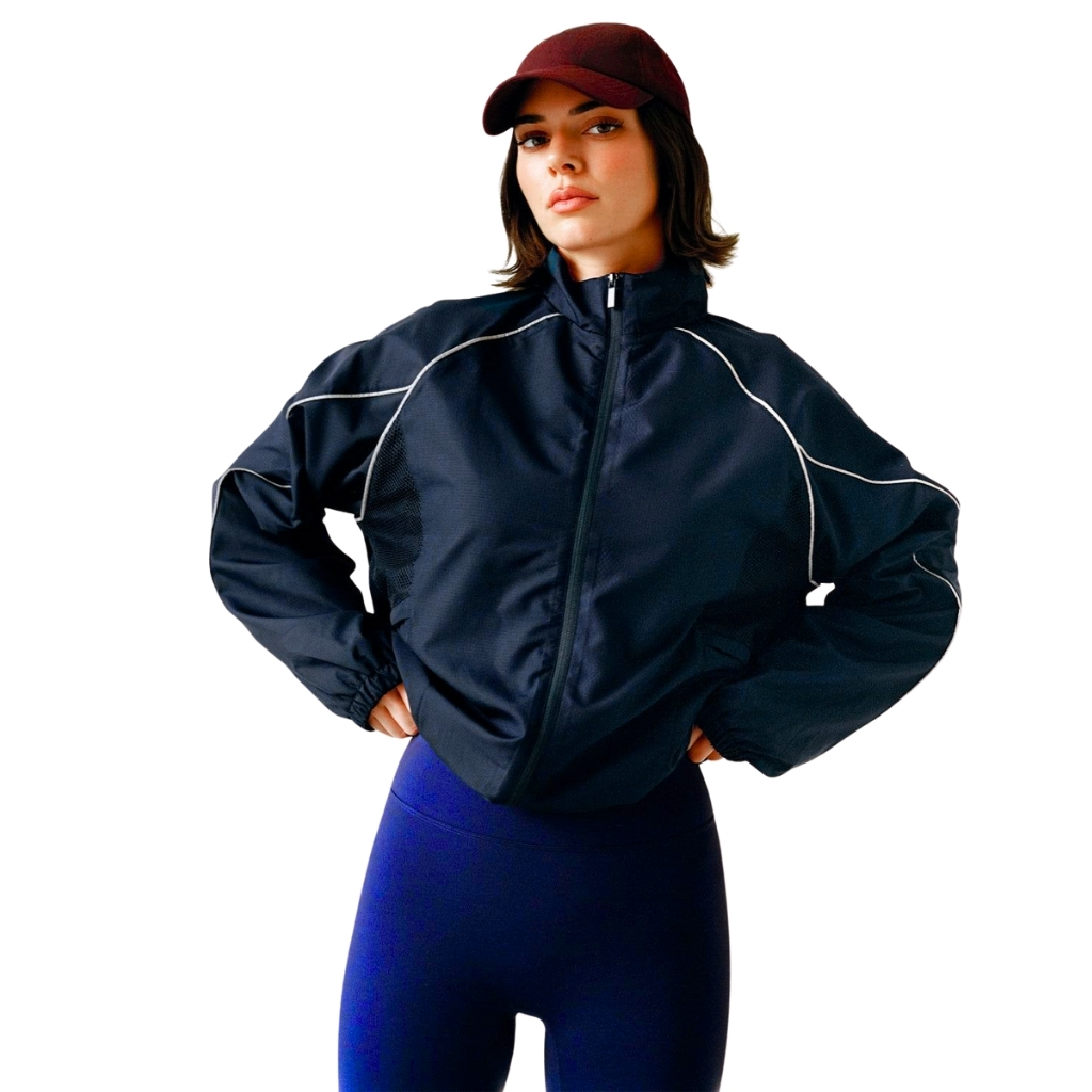 A person model wearing a dark navy windbreaker jacket with white piping, maroon cap, and blue leggings, standing with hands on hips.