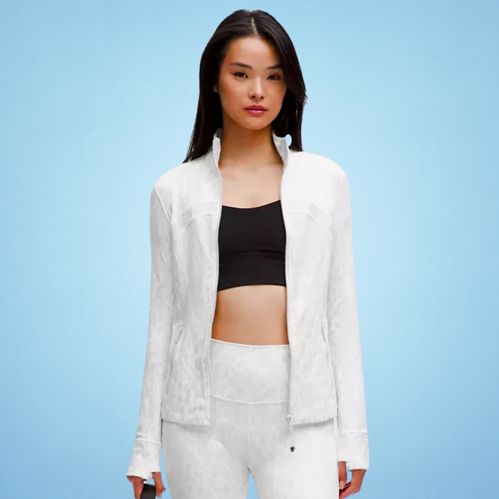 A young woman stands against a light blue background, wearing a white zip-up jacket over a black sports bra and white leggings. She faces the camera with a neutral expression.