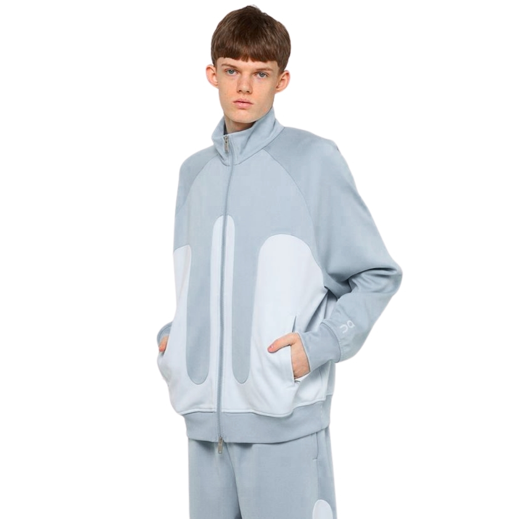 A young man wearing a light blue oversized zip-up jacket with large pale oval panels on the front, matching light blue pants, standing against a white background.