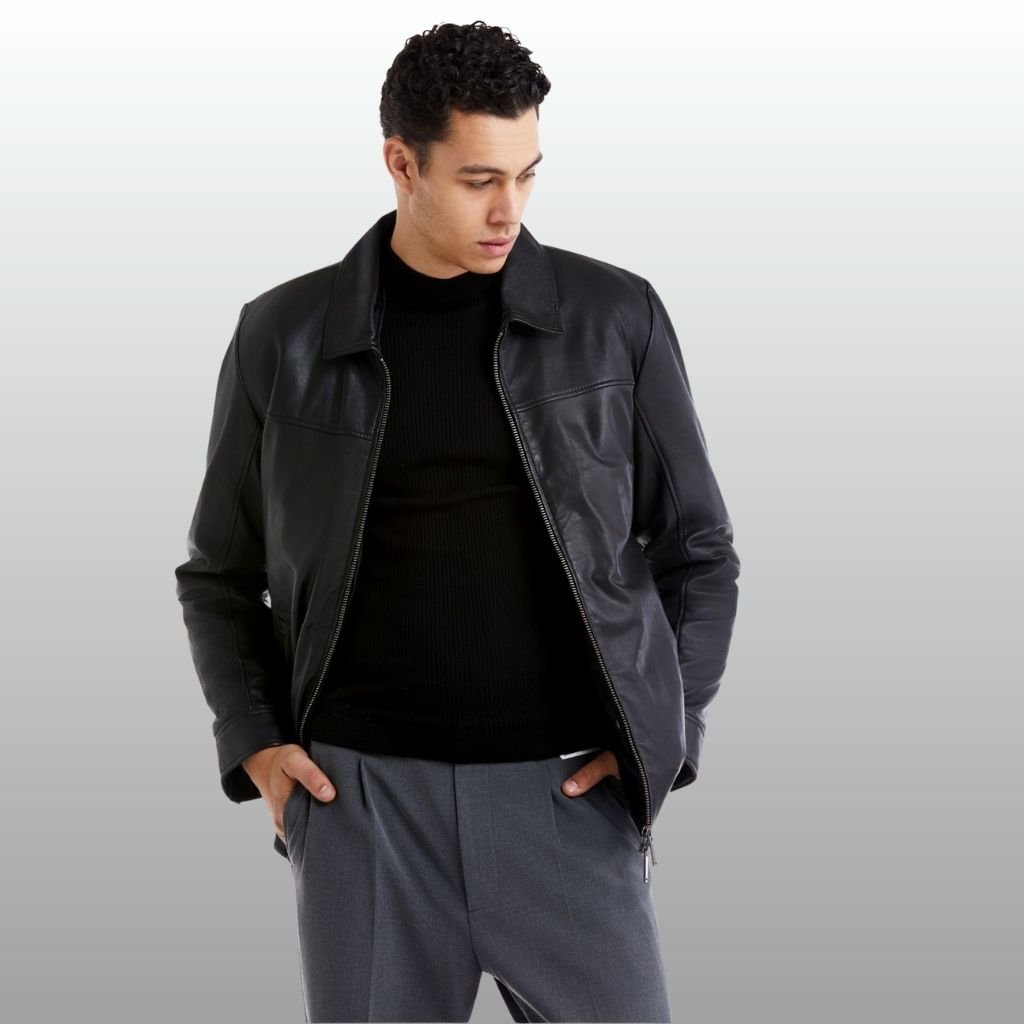 A stylish man wearing a black leather jacket over a black knit sweater and gray trousers, posed with hands in pockets against a soft gradient background.