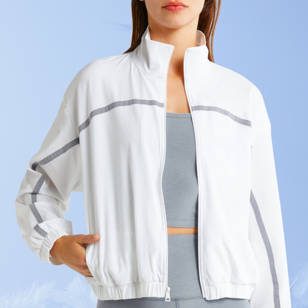 A person wearing a white zip-up athletic jacket with gray piping, standing against a light blue background. The jacket is slightly cropped, with the zipper partly open to reveal a light blue sports bra/top underneath.
