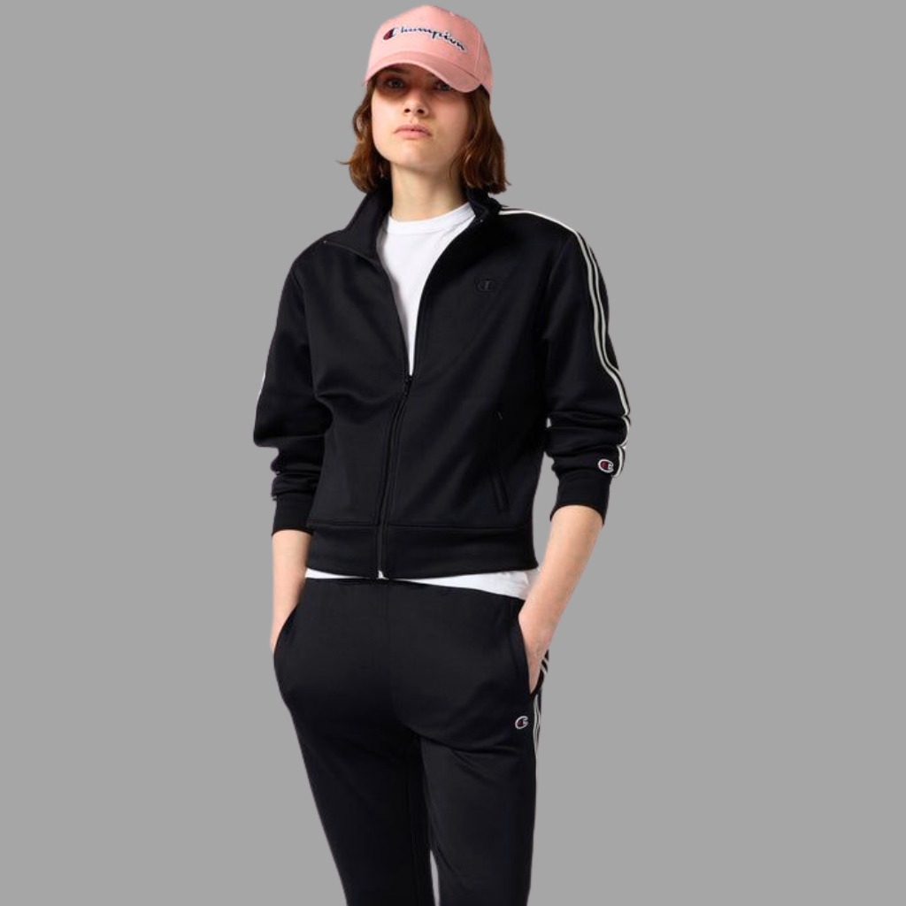 A person wearing a black zip-up tracksuit with white stripes down the sleeves, white undershirt, and a pink cap, standing against a gray background.
