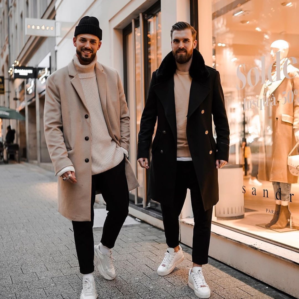 Two stylish men walking down a city street in front of a shop window; both wear long coats, light sweaters, black pants, white sneakers, and beanies. The scene has a chic urban vibe.