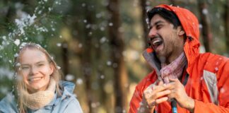 Travel Rain Jackets: Lightweight, Packable & Weatherproof A joyful winter scene of a young woman and a man outdoors in a snowy forest. The woman, with blonde hair, wears a light blue jacket and a beige scarf, smiling. The man, with dark hair and a beard, wears an orange hooded jacket and a colorful scarf, laughing. Snow is on their hair and clothes, and blurred trees and falling snow form the background.