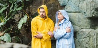 Trench Style Raincoat: Timeless Waterproof Fashion Guides A man in a bright yellow hooded raincoat stands beside a woman in a light blue hooded raincoat. They look in opposite directions, with a leafy backdrop on the left and a textured rock wall on the right.