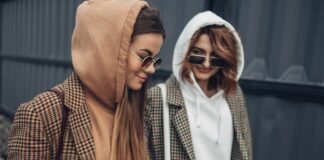 Types of Hoodie Jacket: Styles, Fits, and Trends Guide Two women stand outdoors wearing hoodies and plaid jackets. The woman on the left has long brown hair, a brown hoodie, and sunglasses; the woman on the right has short auburn hair, a white hoodie, sunglasses, and both wear plaid jackets. They smile softly as they look downward, with a gray wall behind them.