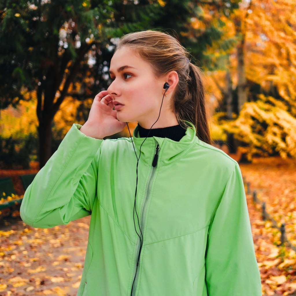 A young woman in a bright green windbreaker headphones in, standing outdoors on a fall day with orange and yellow leaves in the background.