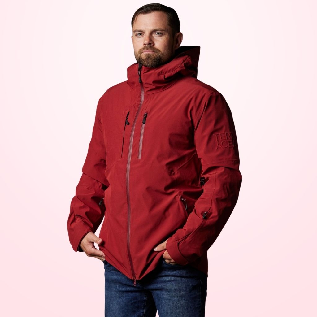 A man wearing a red storm jacket stands against a light pink background, hands in pockets, looking at the camera.