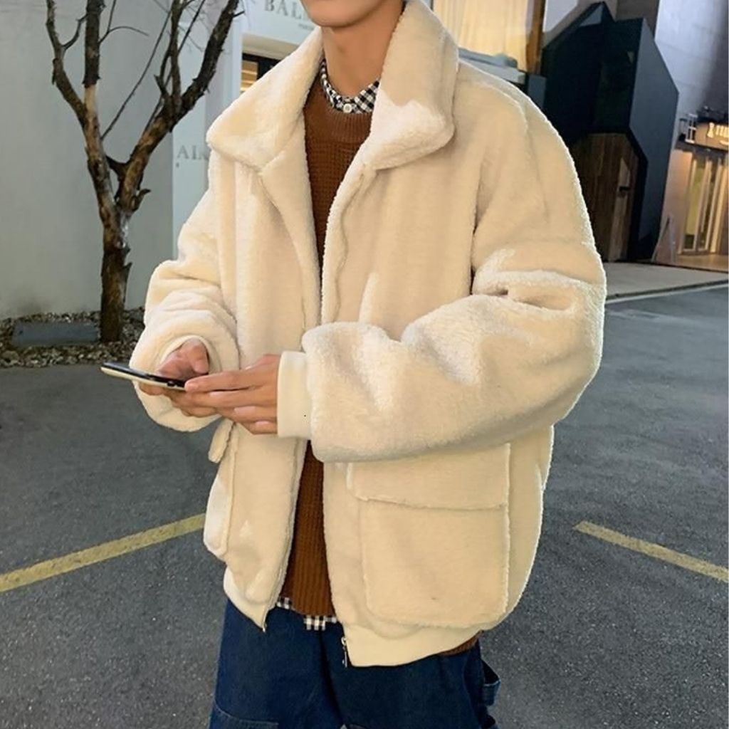 A person stands outdoors wearing a soft, cream-colored teddy jacket with a chunky collar. They are using a smartphone, and a brown knit sweater with a checked shirt peeks out from underneath. The look is completed with dark blue jeans. The setting appears to be a paved street with some trees and storefronts in the background.Plush Jacket for Men