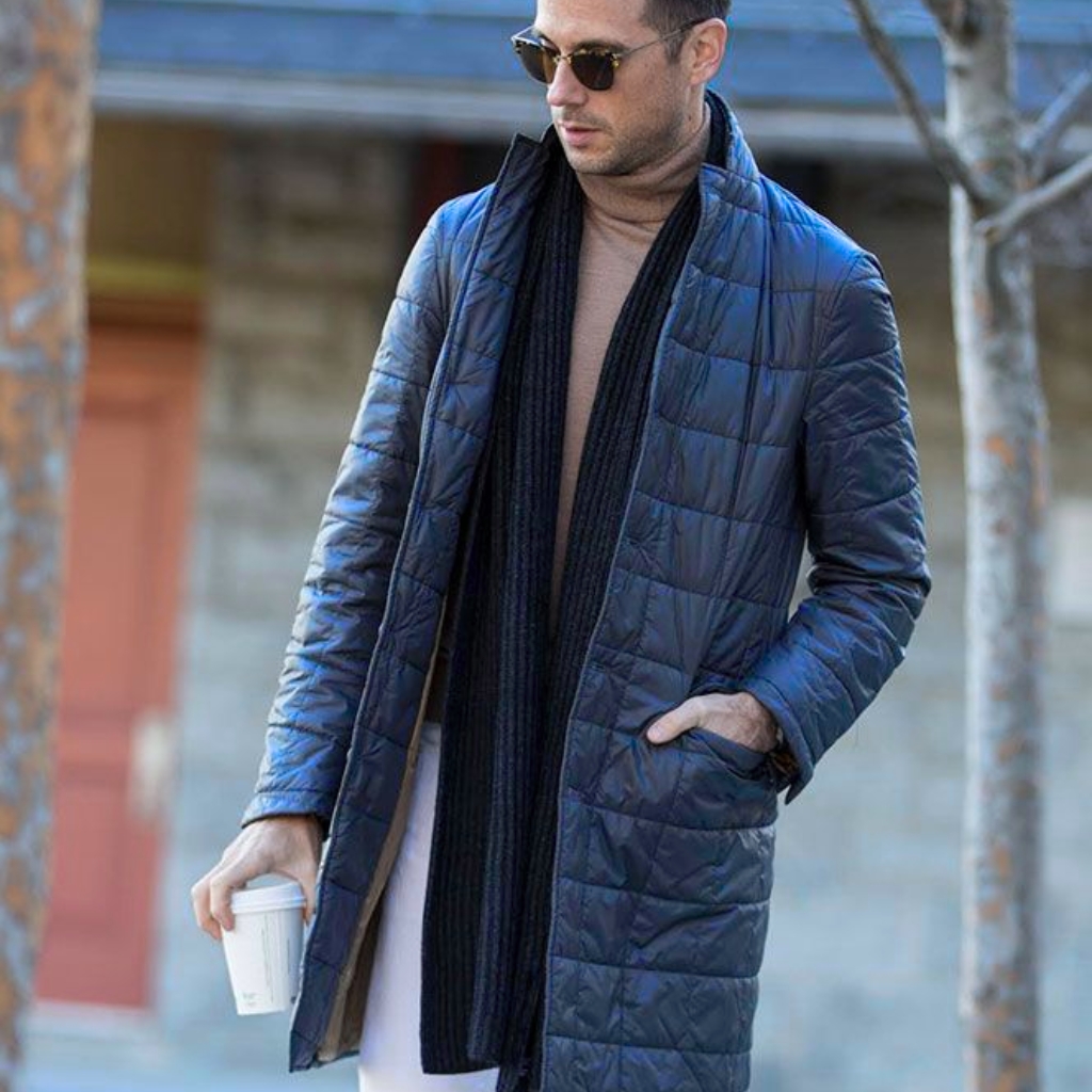  A stylish man wearing a knee-length, quilted blue coat, tan turtleneck, dark scarf, sunglasses, and holding a takeaway coffee cup while walking outdoors.