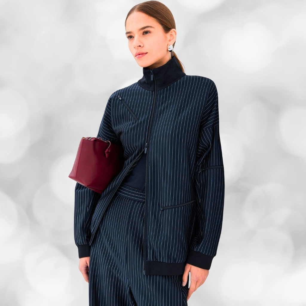 A woman wearing a dark blue pinstripe zip-up jacket with a high collar and matching pinstripe pants. She carries a small burgundy clutch under her left arm and wears white pearl earrings against a soft, blurred light-gray background.