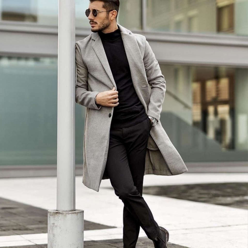 A man wearing a light gray overcoat, black turtleneck, and black pants stands beside a street lamp, posing with one hand on the coat.