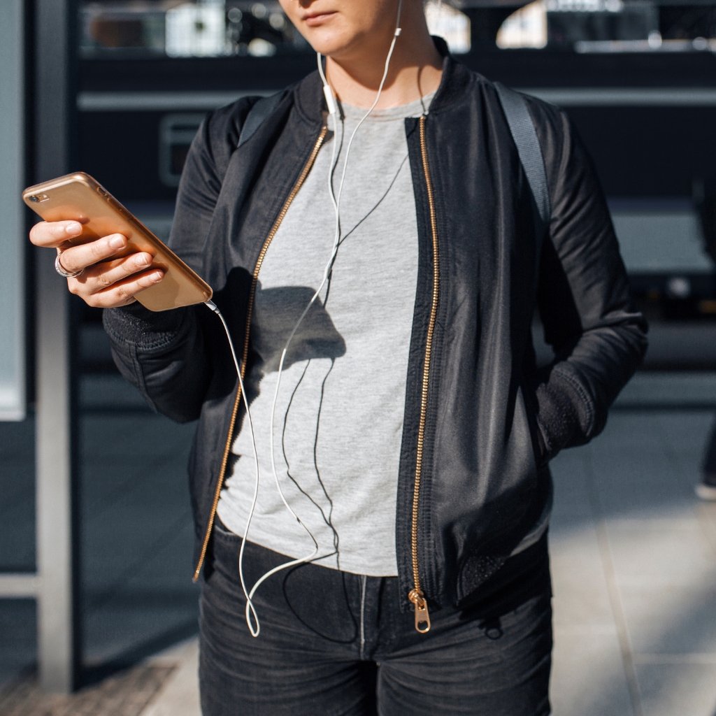 A person stands outdoors, holding a rose-gold smartphone in one hand while wearing white earphones. They’re dressed in a light gray T-shirt, a black jacket with gold zippers, and dark jeans, with a backpack on their shoulders.