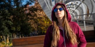 Zip-Up Hoodie: Stylish, comfort and Perfect for Every Season A young woman with long brown hair wearing a maroon zip-up hoodie and blue reflective sunglasses stands outdoors, looking up and to the left, with autumn trees and a metal bridge structure in the background.