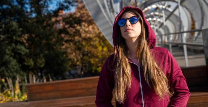 A young woman with long brown hair wearing a maroon zip-up hoodie and blue reflective sunglasses stands outdoors, looking up and to the left, with autumn trees and a metal bridge structure in the background.
