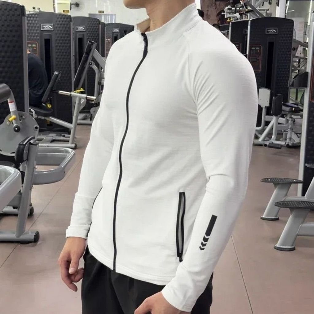 A man wearing a white athletic jacket with a black front zipper, standing in a gym with various exercise machines in the background.Men's Gym Jacket