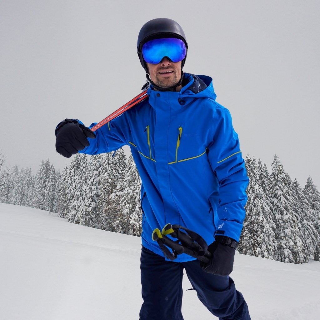 Ski Jacket: Winter Protection, Warmth, & Performance 5 A man wearing a bright blue ski jacket, black helmet, and reflective blue goggles stands in a snowy landscape with snow-covered trees in the background. He holds a red strap over his shoulder and wears black gloves.