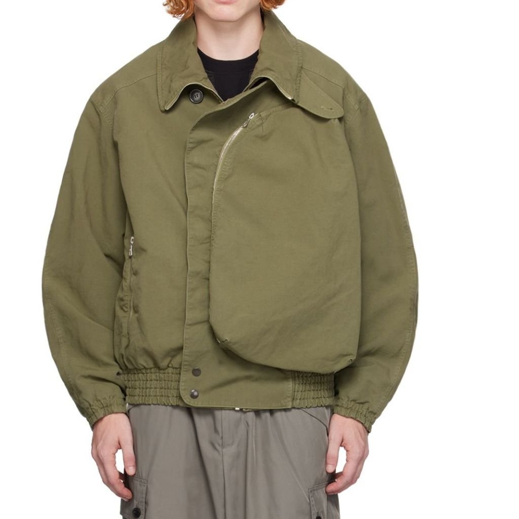  A person wearing an olive-green oversized nylon jacket with a high-low asymmetrical zipper, elasticated cuffs, and a cinched waistband. The jacket features a large, overlapping front panel and a side zipper detail.