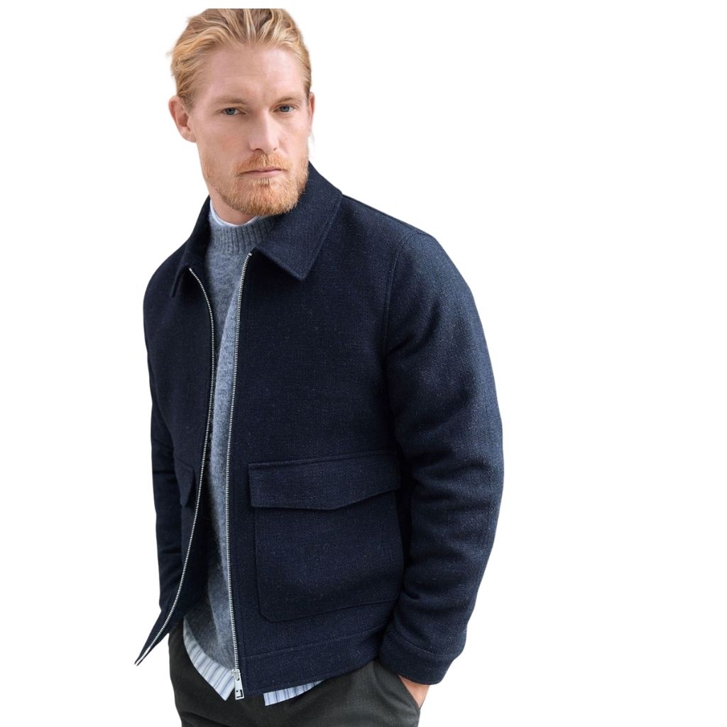 A man wearing a navy blue zip-up jacket with a standing collar, layered over a light gray knit sweater; he has blond hair and a light beard, and he stands against a plain white background.