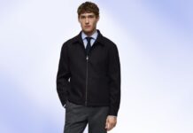 Office Jacket: The Best Blend of Style and Comfort Young man wearing a black zip-front jacket over a light blue shirt and dark tie, standing against a soft blue background.