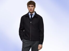 Young man wearing a black zip-front jacket over a light blue shirt and dark tie, standing against a soft blue background.