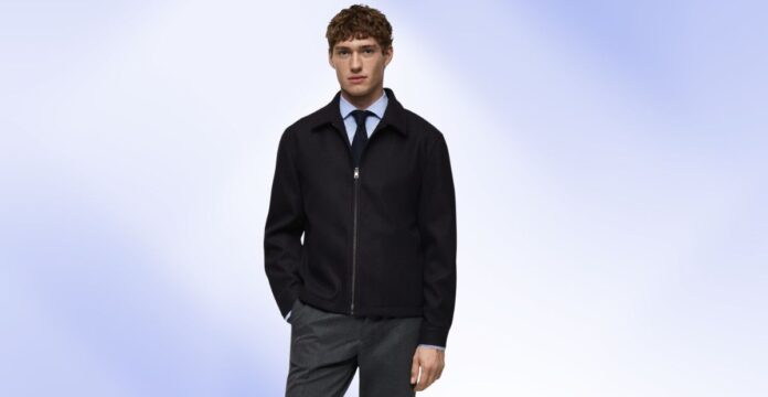 Young man wearing a black zip-front jacket over a light blue shirt and dark tie, standing against a soft blue background.