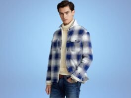A stylish young man stands against a light blue background, wearing a blue-and-white plaid jacket over a cream turtleneck sweater and jeans.