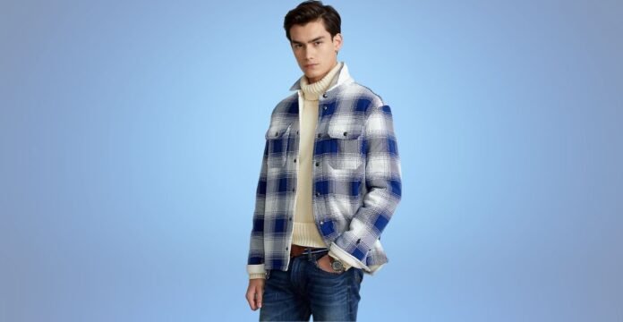 A stylish young man stands against a light blue background, wearing a blue-and-white plaid jacket over a cream turtleneck sweater and jeans.
