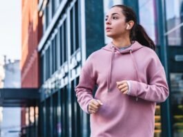 A young woman jogging outdoors in an urban area, wearing a pink hoodie and wireless earbuds, with glass-fronted buildings in the background.