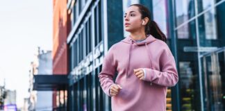 Athletic Hoodie: Comfort, Style & Performance Wear A young woman jogging outdoors in an urban area, wearing a pink hoodie and wireless earbuds, with glass-fronted buildings in the background.