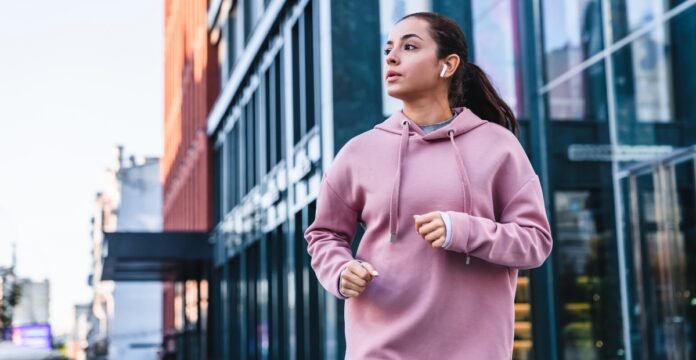 A young woman jogging outdoors in an urban area, wearing a pink hoodie and wireless earbuds, with glass-fronted buildings in the background.
