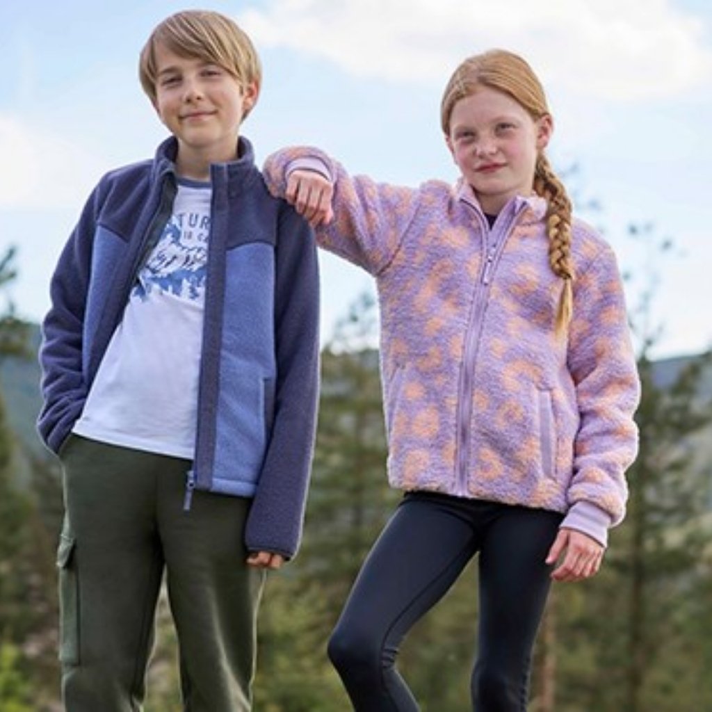 A young boy and girl stand outdoors side by side, with a backdrop of trees and a partly cloudy sky. The boy has blonde hair and wears a blue-purple fleece jacket over a white graphic t-shirt and dark green cargo pants. The girl, with red braided hair, rests her arm on the boy’s shoulder and wears a lavender fleece jacket with an orange geometric pattern and black leggings.