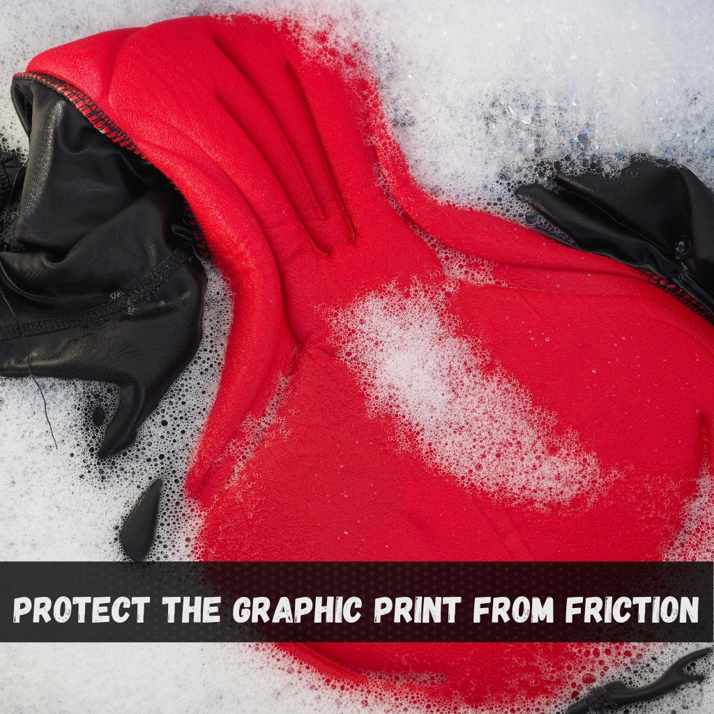 Graphic Full-Zip Hoodies: Stylish Comfort for Every Occasion 4 A red and black hoodie being washed in soapy water, with white suds surrounding it; a bold black banner at the bottom reads "PROTECT THE GRAPHIC PRINT FROM FRICTION."