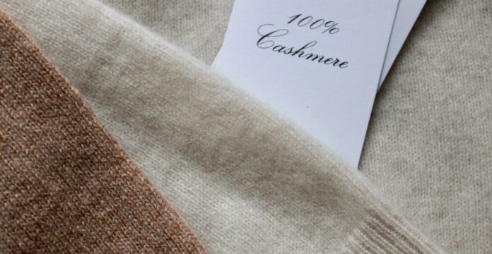 Cashmere Blended Fabric Cashmere Blended Fabric Close-up view of stacked cashmere fabrics in brown and beige tones, with a white tag reading '100% Cashmere' partially visible.