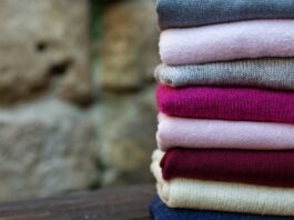 Pure Cashmere Wool Fabric A stack of folded sweaters in various colors (gray, pink, fuchsia, burgundy, cream, and blue) sits on a dark wooden surface with a blurred stone wall in the background.