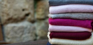Types of Pure Cashmere Wool Fabric Explained Pure Cashmere Wool Fabric A stack of folded sweaters in various colors (gray, pink, fuchsia, burgundy, cream, and blue) sits on a dark wooden surface with a blurred stone wall in the background.