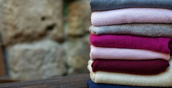 Pure Cashmere Wool Fabric A stack of folded sweaters in various colors (gray, pink, fuchsia, burgundy, cream, and blue) sits on a dark wooden surface with a blurred stone wall in the background.