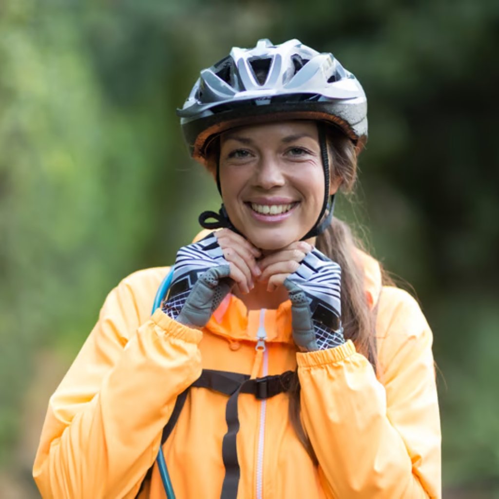 Ultimate Bicycle Rain Jacket for All-Weather Cycling Gear 6 A smiling woman wearing a bicycle helmet and an orange jacket adjusts the strap of her helmet while wearing gloves; a blurred forest background behind her.