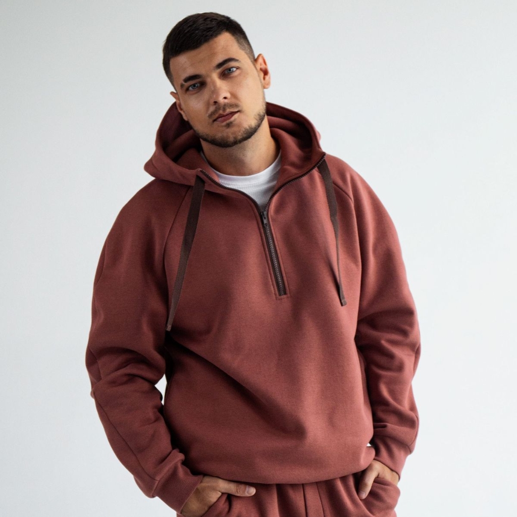 Streetwear Hoodie: Trendy Urban Style for Every Wardrobe 2 A man wearing a terracotta hoodie and matching sweatpants stands against a white background, looking at the camera with a neutral expression. He has short dark hair and a light beard; his hands are in his pockets; the hoodie is half-zip with a white shirt underneath. streetwear hoodie
