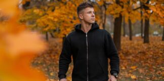 Classic Full-Zip Hoodie: Timeless Style & Everyday Comfort A man wearing a black full-zip hoodie stands outdoors in an autumn setting, with orange and yellow leaves on the trees and ground.