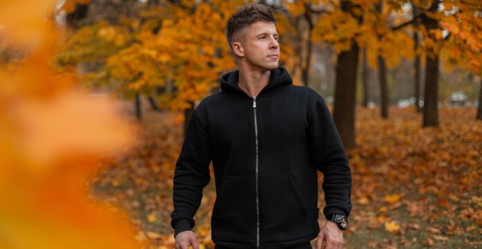 A man wearing a black full-zip hoodie stands outdoors in an autumn setting, with orange and yellow leaves on the trees and ground.