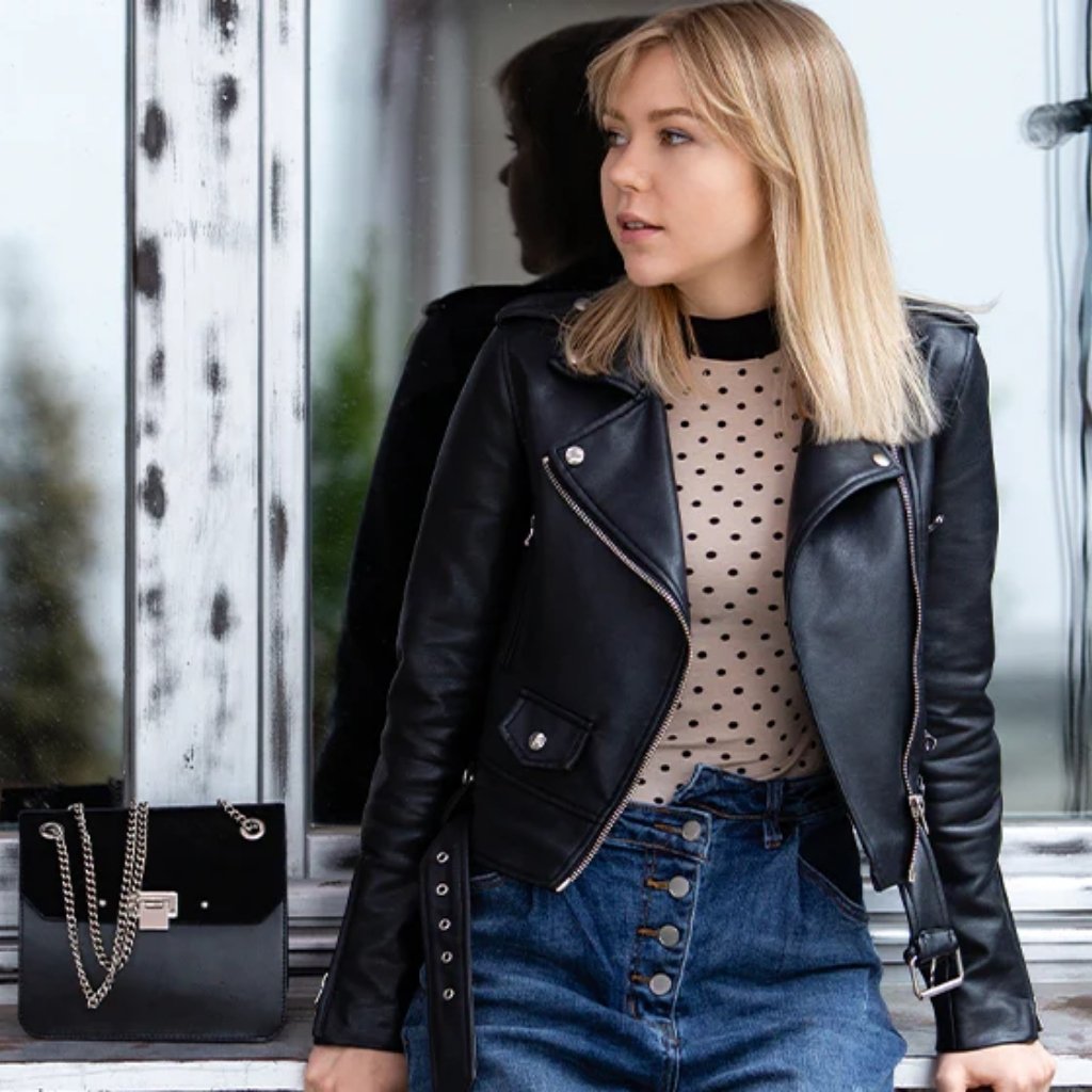 Everyday Leather Jacket – Timeless Comfort and Style for Daily Wear 7 A young woman with blonde hair wearing a black leather jacket, beige polka-dot top, and blue jeans stands beside a window. Her reflection is visible in the glass, and a black purse sits on the ledge beside her.