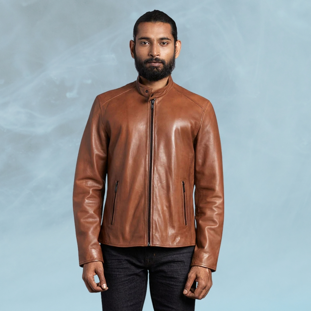 Cognac Leather Jacket Designed for Everyday Style and Comfort 3 A man with a neatly groomed beard and dark hair pulled back stands facing the camera against a hazy blue background. He wears a brown leather jacket zipped up over a dark shirt and dark jeans.