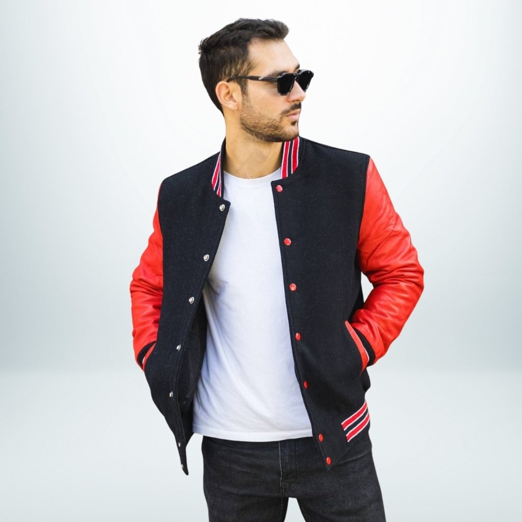 A man wearing a black varsity jacket with red leather sleeves and a white t-shirt, sunglasses, and dark jeans stands with hands in pockets, looking to the side against a pale background.