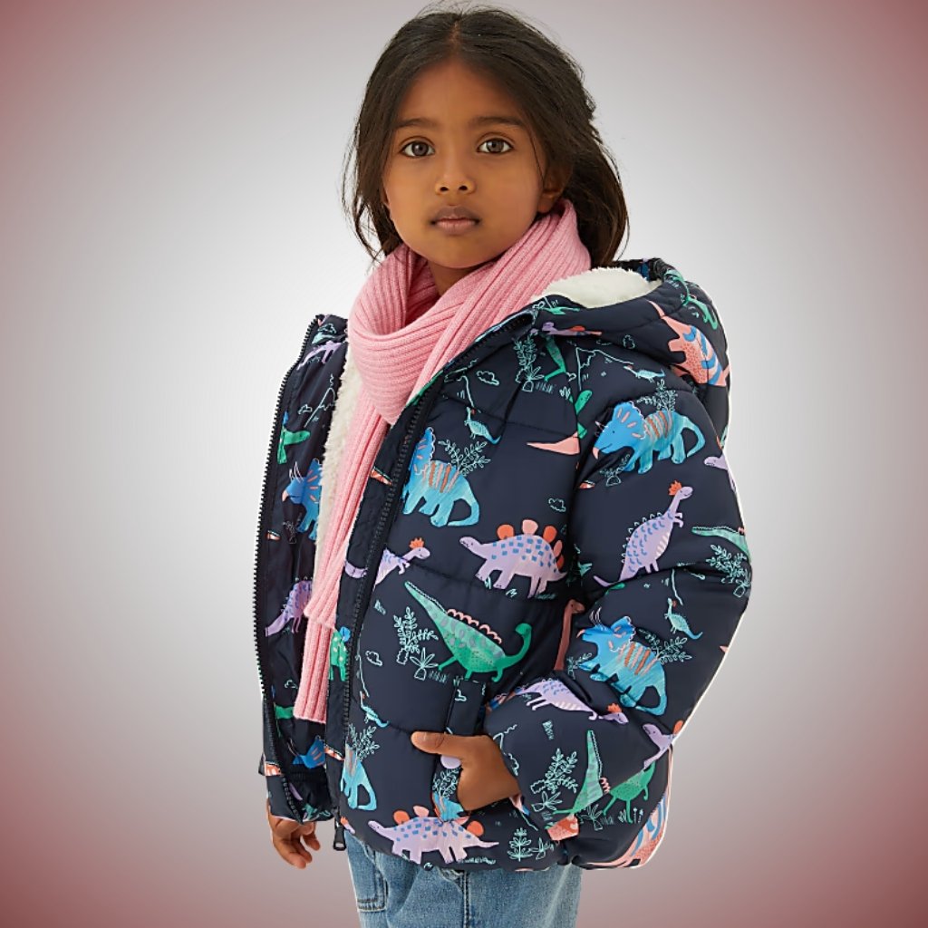 A young girl wearing a navy blue puffer jacket with colorful dinosaur patterns, a light pink scarf, and light blue jeans. She stands against a soft gray-to-red gradient background, with one hand in her jacket pocket and looking at the camera.